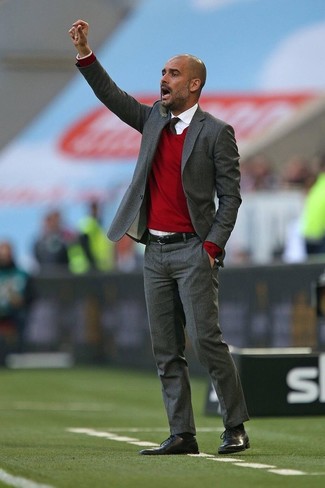 Pep Guardiola trägt grauer Wollanzug, roter Pullover mit einem V-Ausschnitt, weißes Businesshemd, schwarze Leder Oxford Schuhe