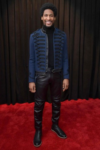 Jon Batiste trägt dunkelblaue bestickte Cabanjacke, schwarzer Rollkragenpullover, schwarze Lederjeans, schwarze Chelsea-Stiefel aus Leder