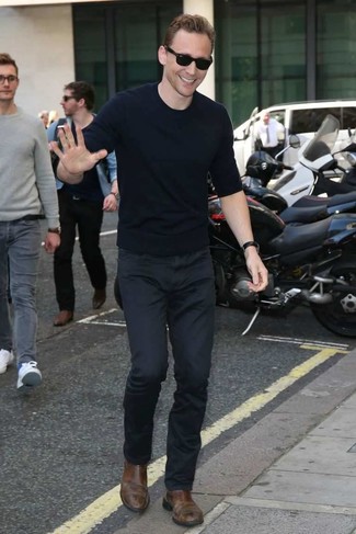 Tom Hiddleston trägt dunkelblauer Pullover mit einem Rundhalsausschnitt, schwarze Chinohose, braune Chelsea-Stiefel aus Leder, schwarze Sonnenbrille