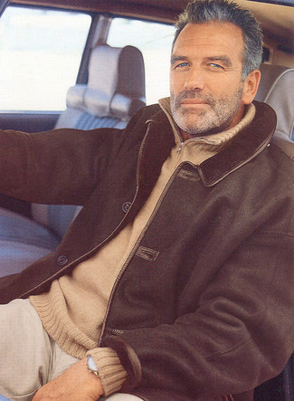 Roy Summersett trägt schwarze Lammfelljacke, beige Pullover mit einem Reißverschluss am Kragen, hellbeige Chinohose