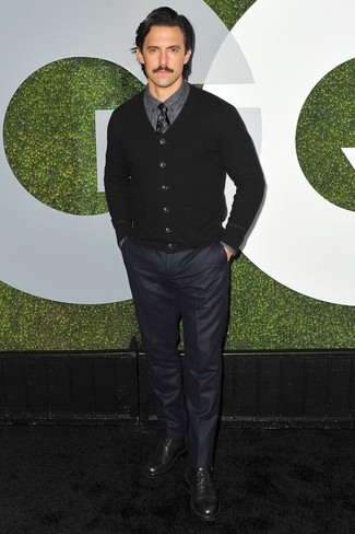Milo Ventimiglia trägt schwarze Strickjacke, dunkelgraues Businesshemd, dunkelblaue Anzughose, schwarze Leder Derby Schuhe