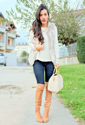 hellbeige Pelzweste, weißer Pullover mit einem Rundhalsausschnitt, dunkelblaue enge Jeans, beige Overknee Stiefel aus Wildleder für Damen